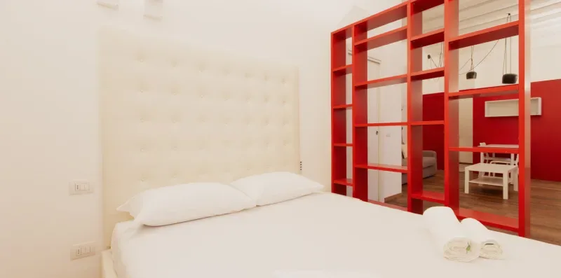 A bedroom with a white bed and a red open shelving divider.