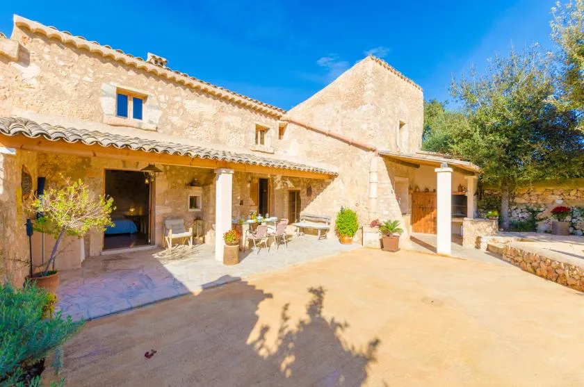 Una casa de piedra da a un patio soleado con asientos a la sombra.
