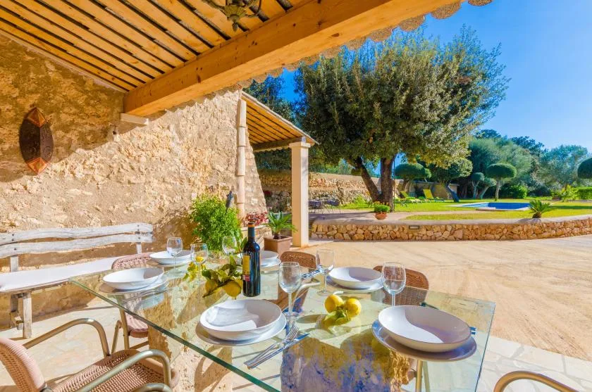 Una mesa de comedor de vidrio está preparada bajo un patio cubierto junto a un muro de piedra.