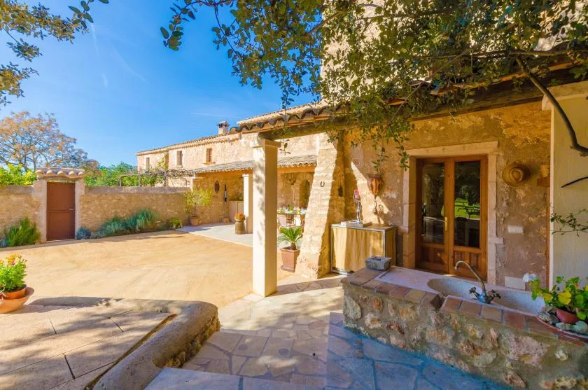 Un patio soleado de piedra con una puerta de madera, una zona de estar sombreada y plantas en macetas.