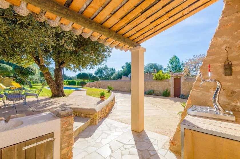 Un patio exterior pavimentado bajo una pérgola de madera, con una barbacoa y asientos de jardín.