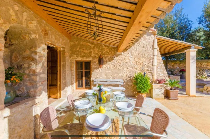 Una zona de comedor en un patio de piedra tiene una mesa de vidrio bajo una pérgola de madera.