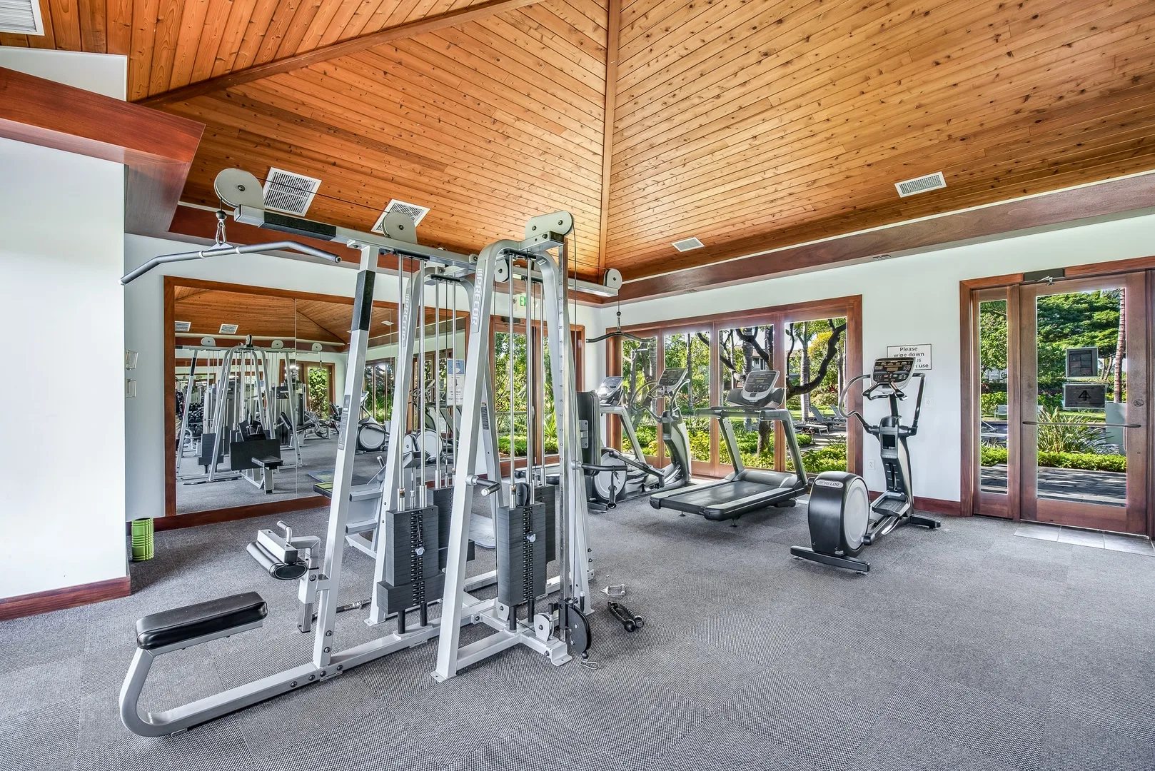 A fitness room with exercise machines, mirrored walls, and windows overlooking greenery.