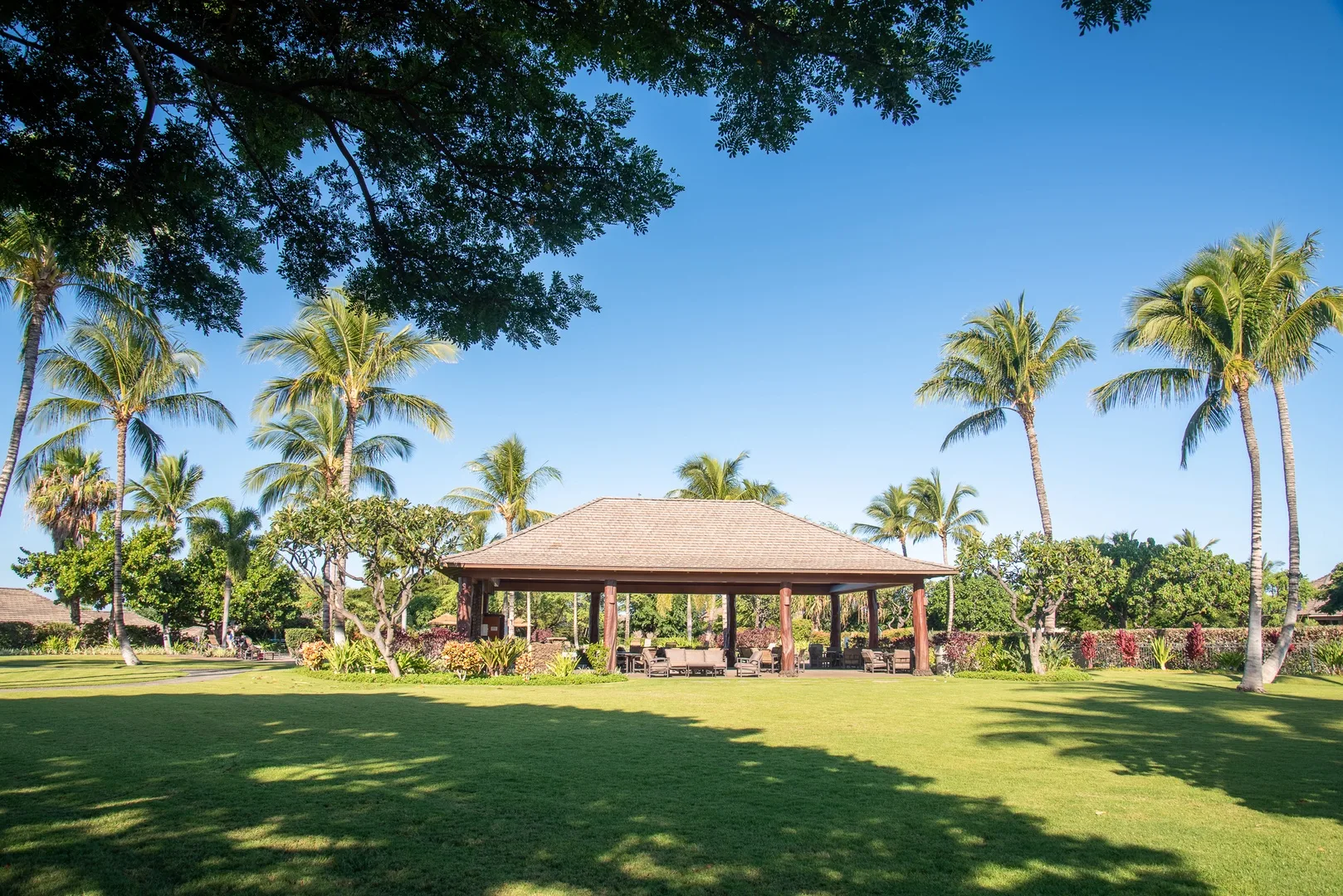 A roofed pavilion stands on a broad green lawn among palm trees.