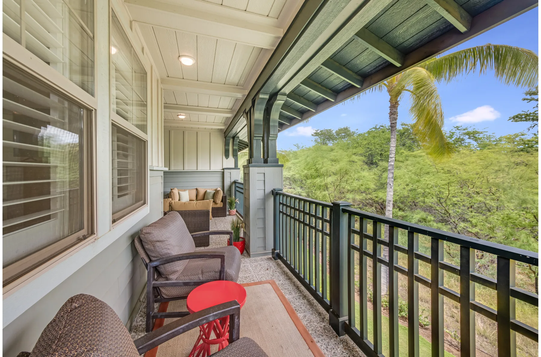 A covered balcony has seating beside a black railing overlooking palm trees and greenery.