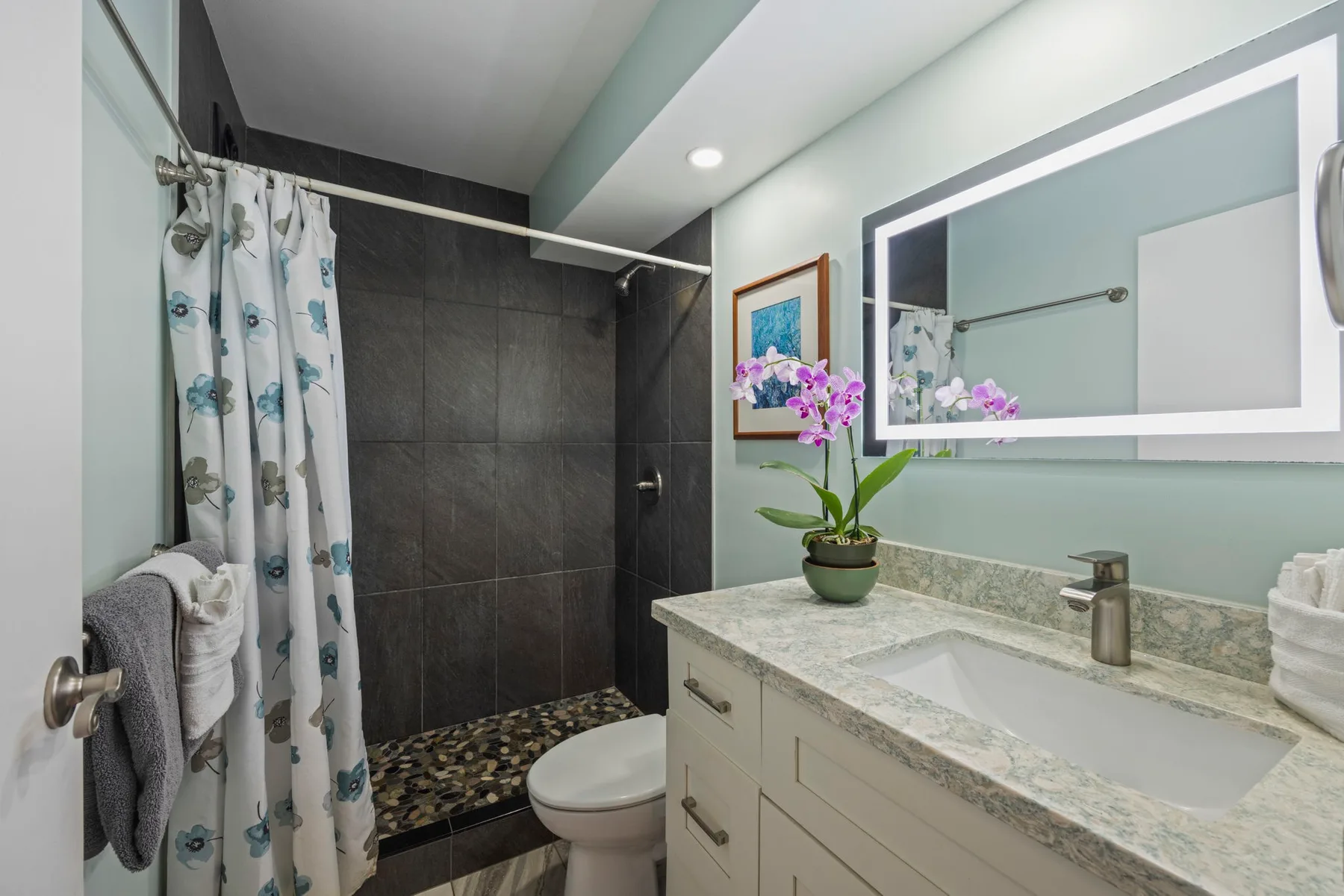 Un baño con una zona de ducha alicatada y un tocador con una planta en flor.