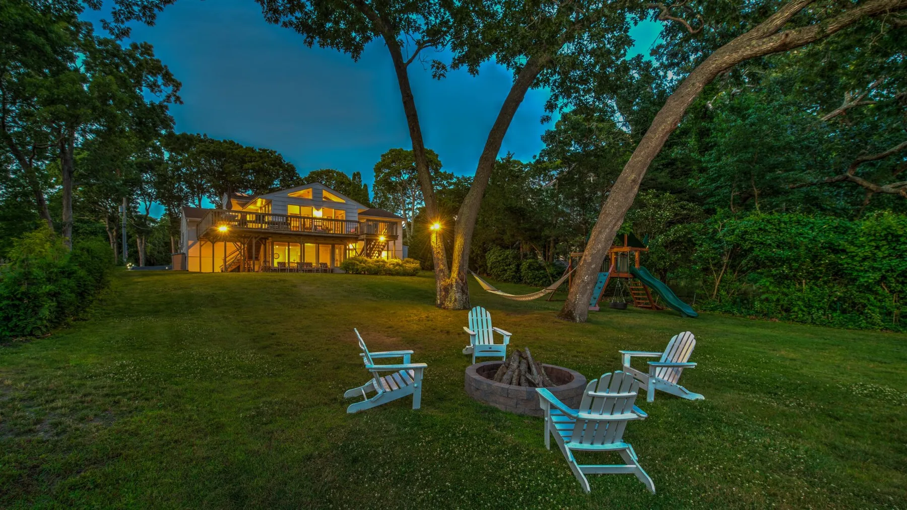 A wooded backyard at dusk features a lit house, lawn seating, and a fire pit.