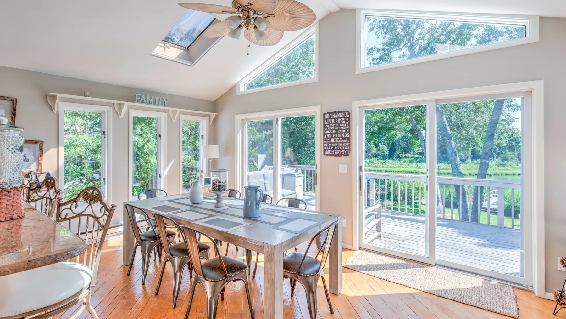 A bright dining area with a long table beside large windows overlooking greenery.