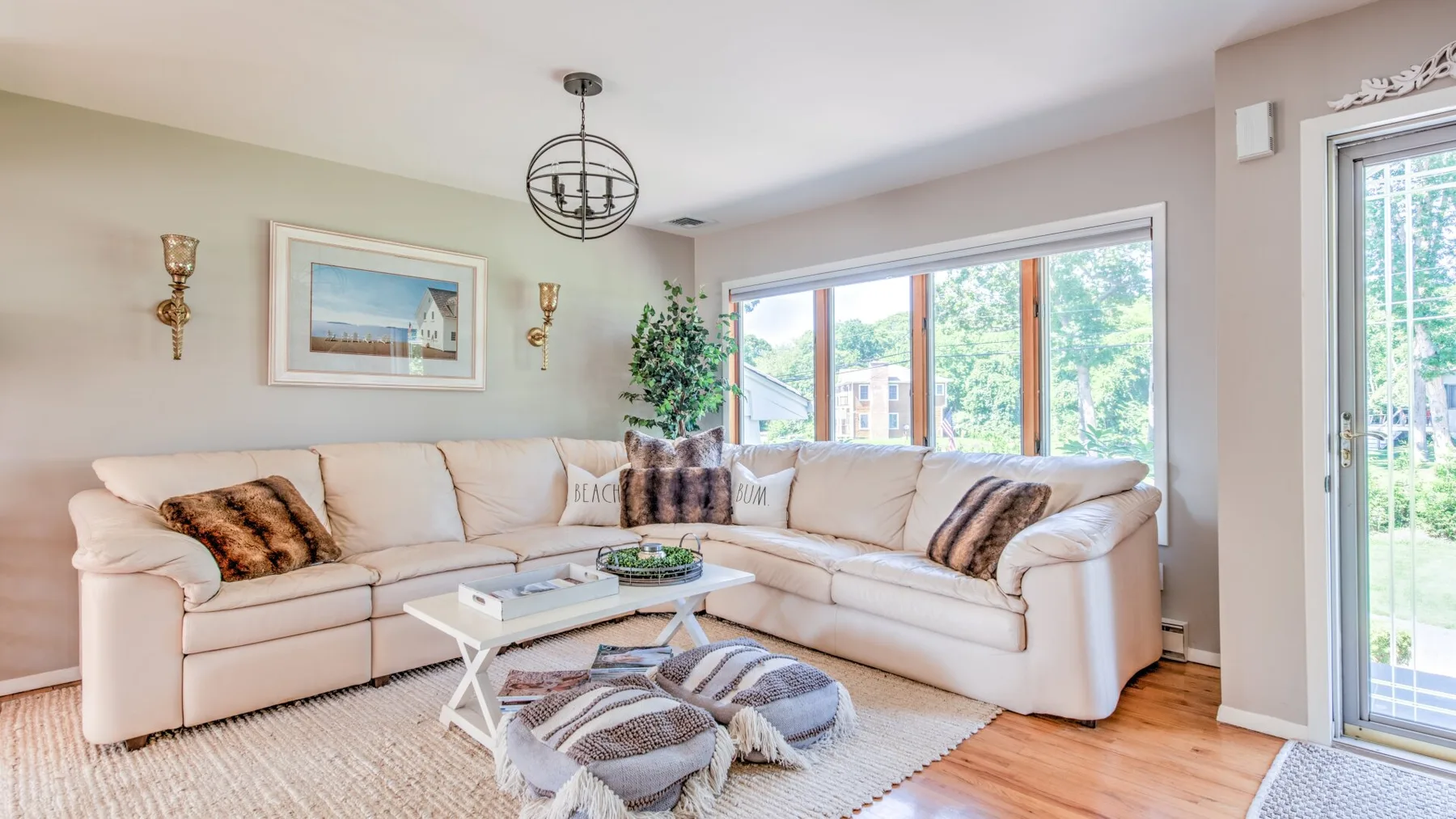 A bright living room has a pale sectional sofa, a coffee table, and large windows.