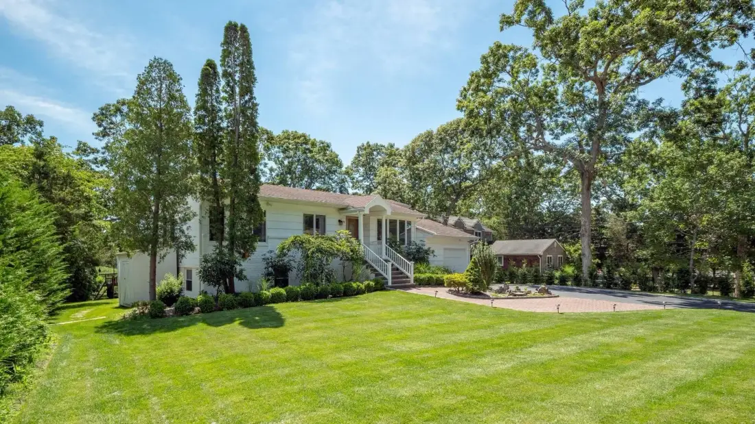 A house sits beside a broad green lawn, with mature trees surrounding the yard.
