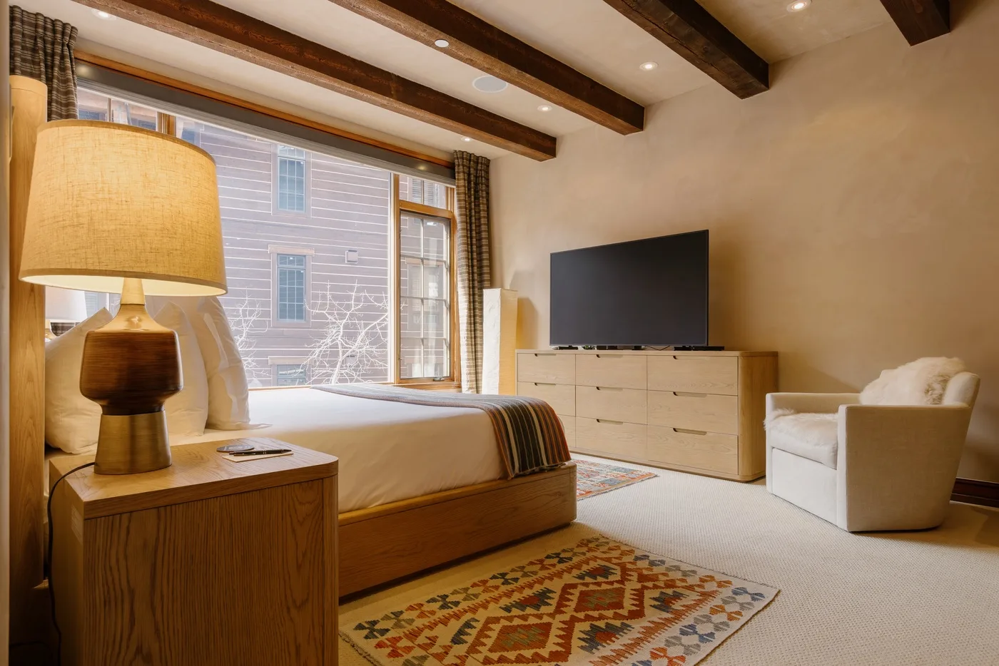 Un dormitorio con cama, cómoda, televisor y sillón junto a una ventana grande.