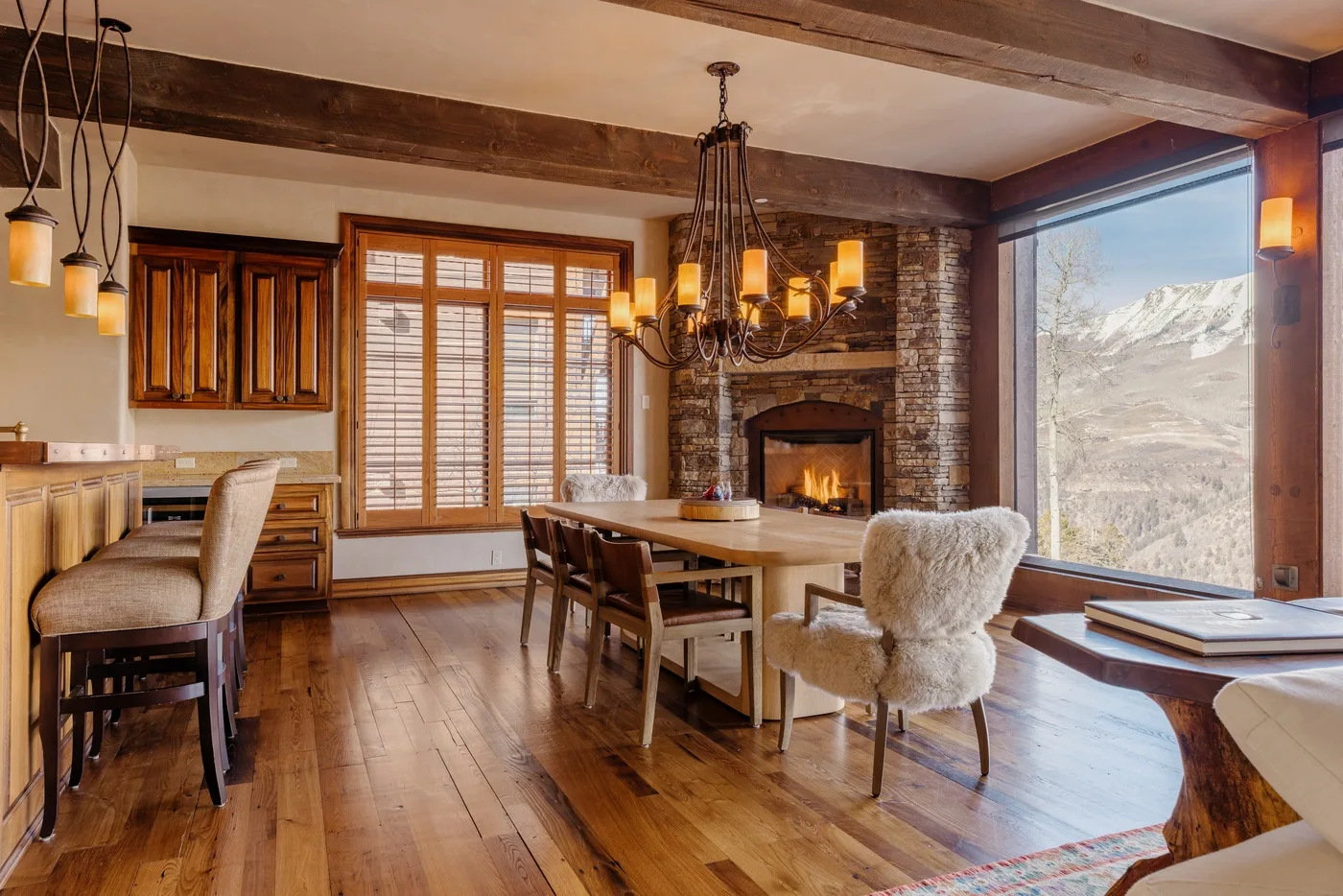 Comedor rústico con mesa de madera, chimenea y vistas a las montañas por una ventana amplia.
