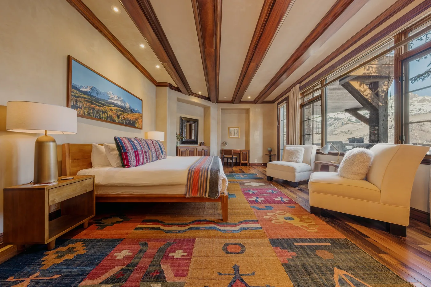 Un dormitorio con una cama grande, alfombras coloridas y asientos junto a amplias ventanas.