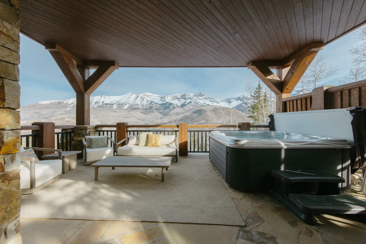 Una zona exterior cubierta con asientos y una bañera de hidromasaje mira hacia montañas nevadas.