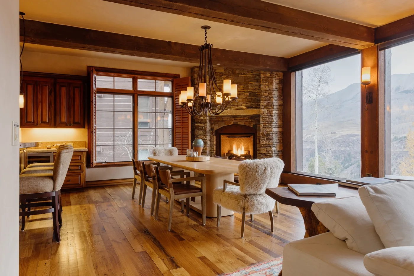 A warm dining and living area features a wooden table, fireplace, and large windows.