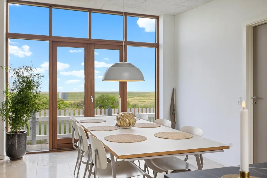 A dining area with a long table and chairs beside large windows overlooking a green landscape.