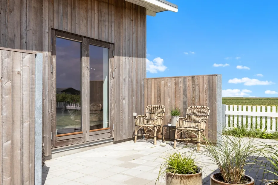 An outdoor patio beside a wooden exterior wall, with wicker seating and potted grasses.