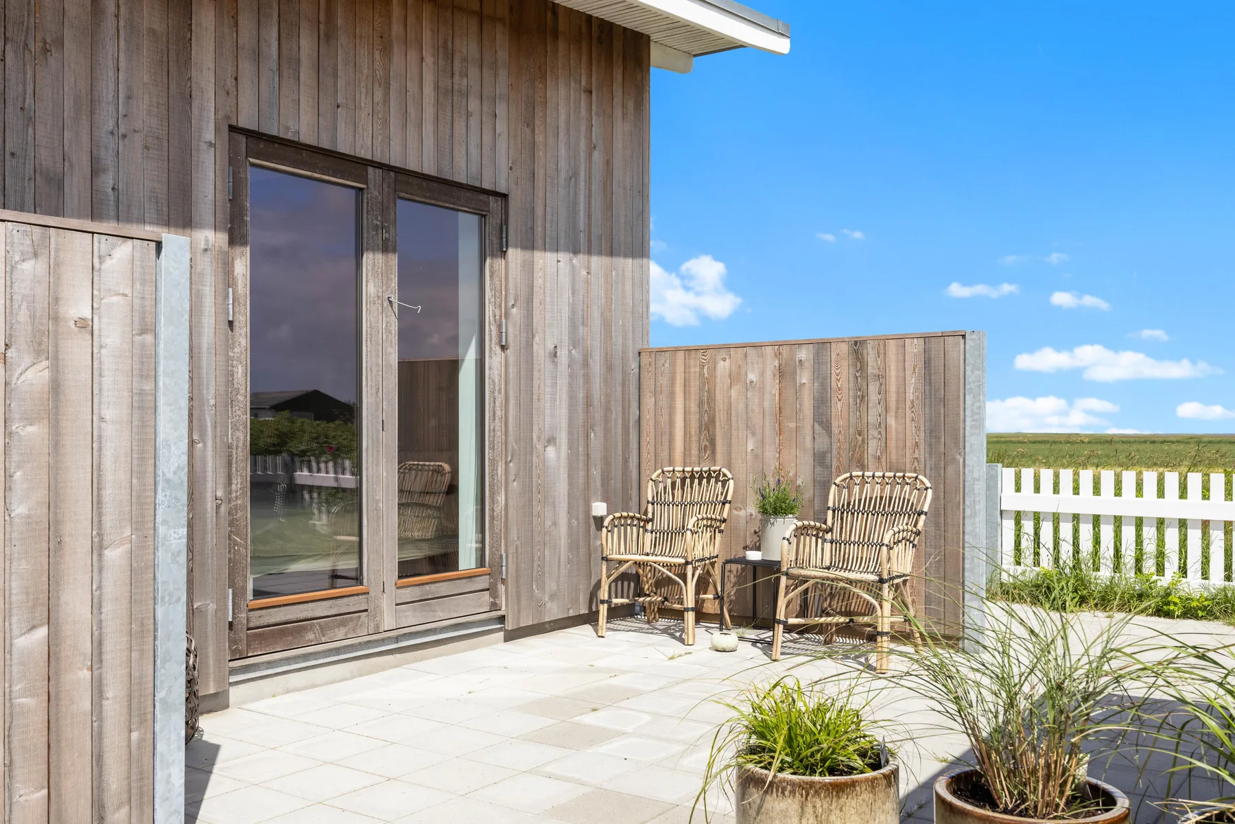 A sunny outdoor patio beside a wooden-sided building, with seating and potted plants.