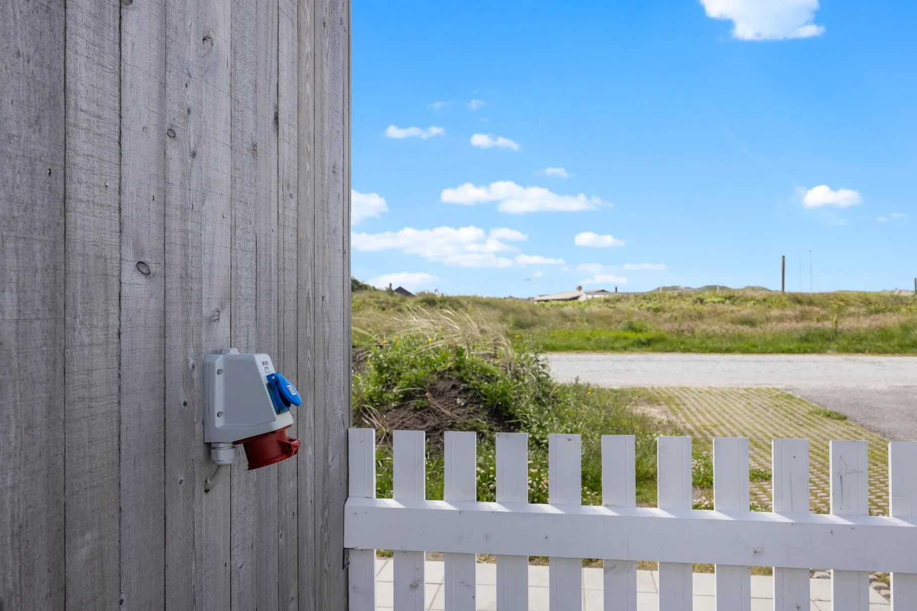 A white gate beside a wooden wall opens onto a sandy path through grassy dunes.