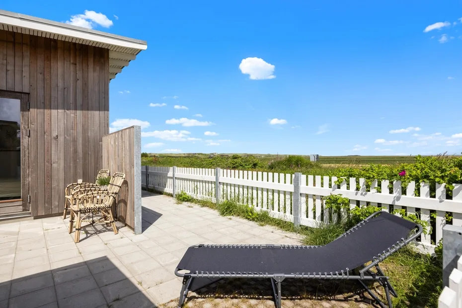 A sunny outdoor patio with a lounge chair, seating, and a white fence.