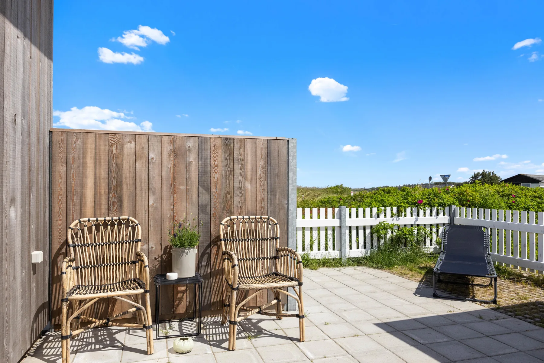 A sunny patio with seating beside a wooden privacy screen and white fence.