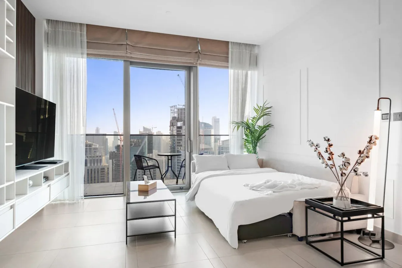 A bright bedroom with a bed, seating, and large windows opening onto a city view.