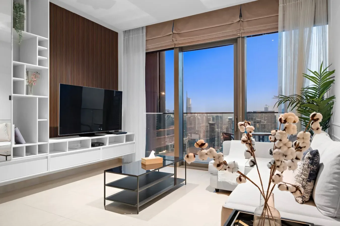 A bright living room with a television, low table, and large windows overlooking the city.