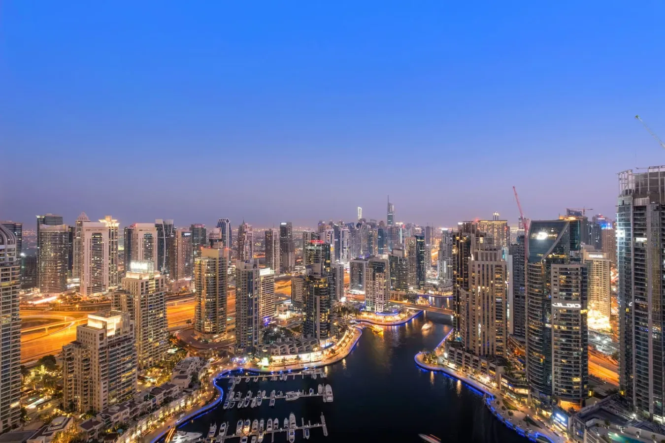 A city skyline at dusk surrounds a marina with boats and illuminated waterfront paths.