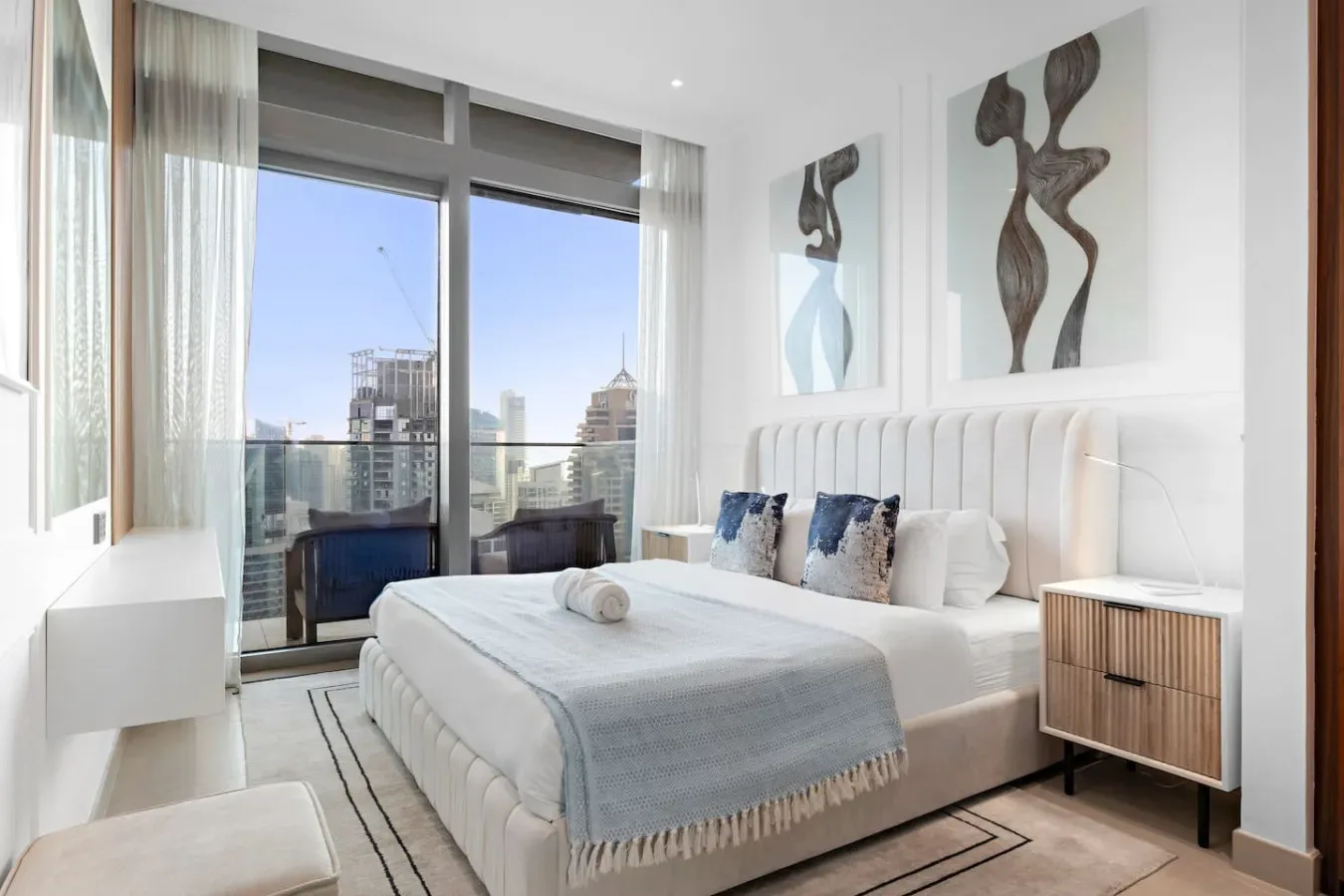 A bright bedroom with a neatly made bed beside large windows overlooking city buildings.