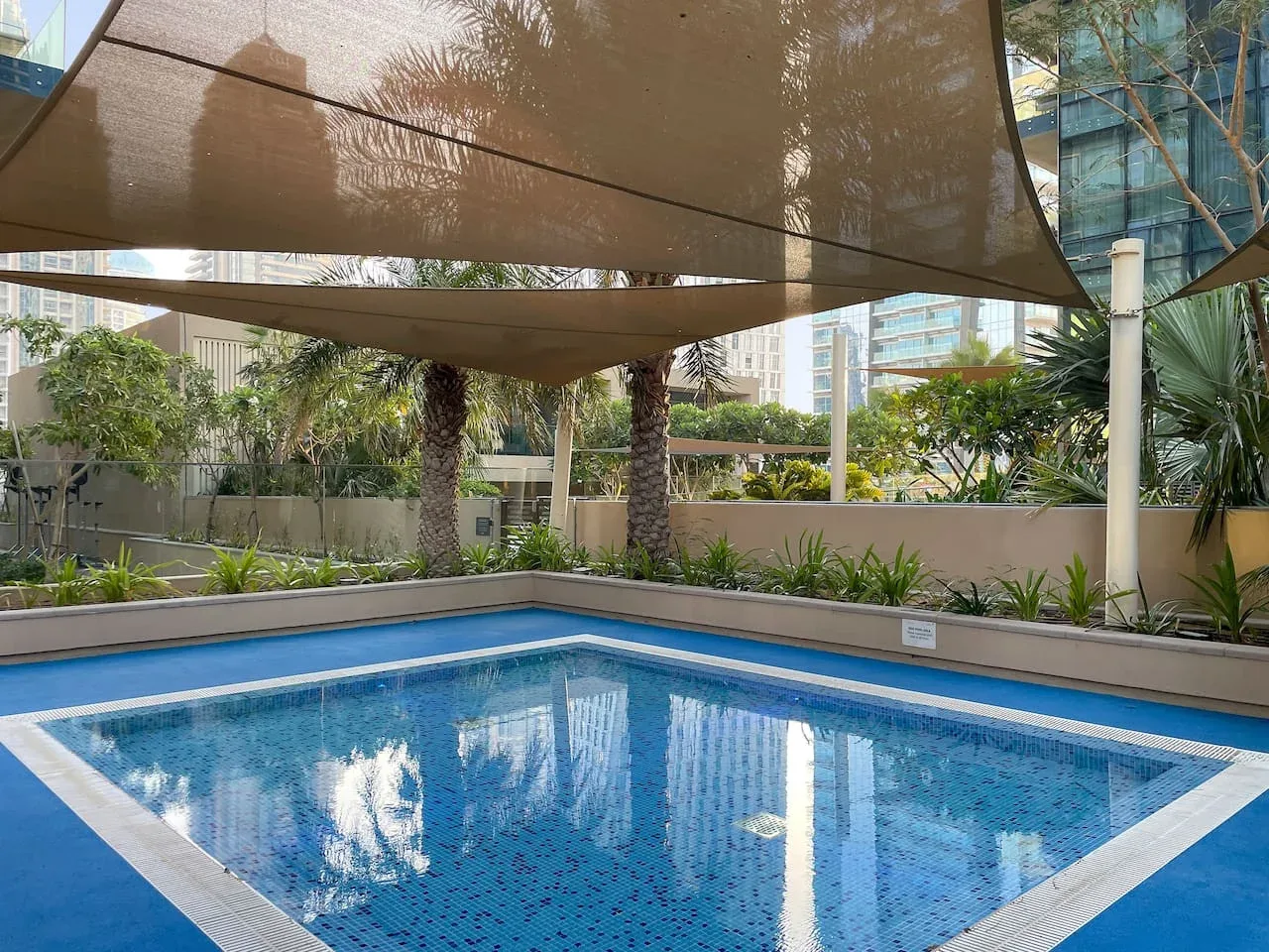 An outdoor swimming pool sits beneath large shade sails, surrounded by planted landscaping.