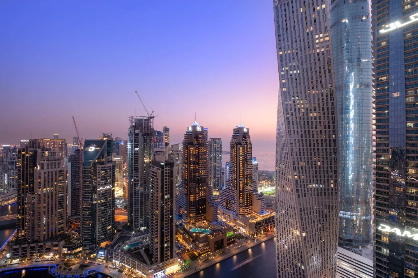 A city skyline at dusk, with illuminated high-rise buildings beside a waterway.
