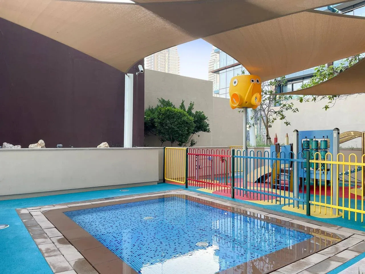 A shallow outdoor pool sits beneath shade sails beside a colorful play area.