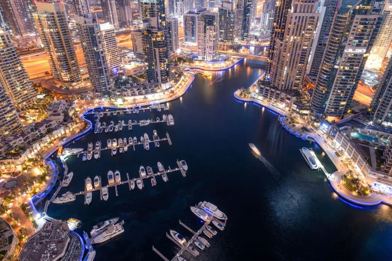 Aerial view of a marina with moored boats and illuminated waterfront buildings at night.