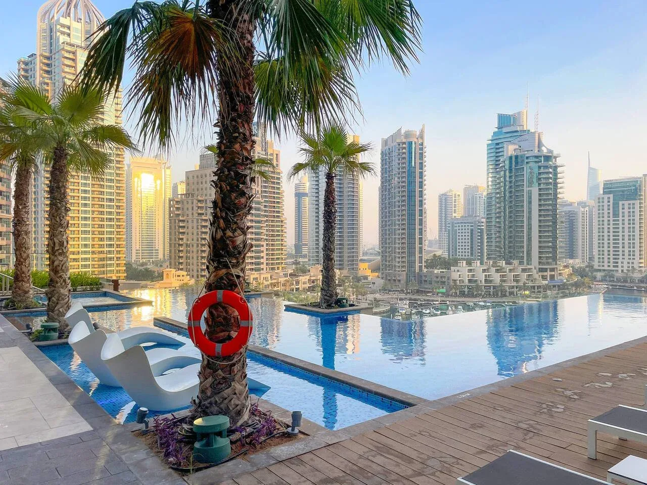 An outdoor pool beside a paved deck, palm trees, and a city skyline.
