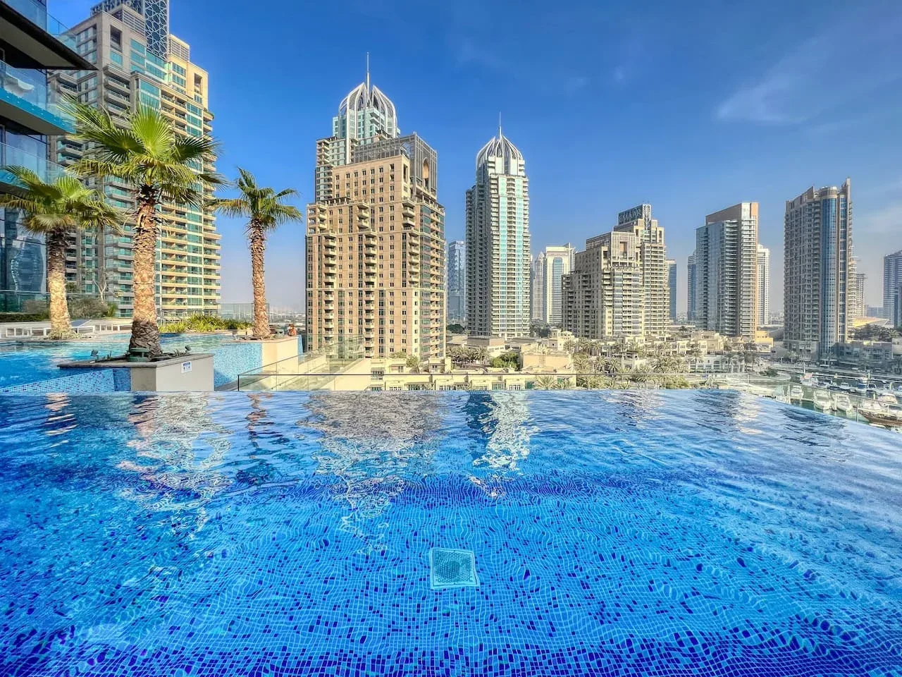 A rooftop swimming pool overlooks a cluster of high-rise buildings under a clear blue sky.
