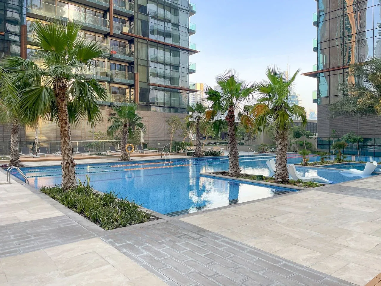Outdoor swimming pool with palm trees between modern buildings.