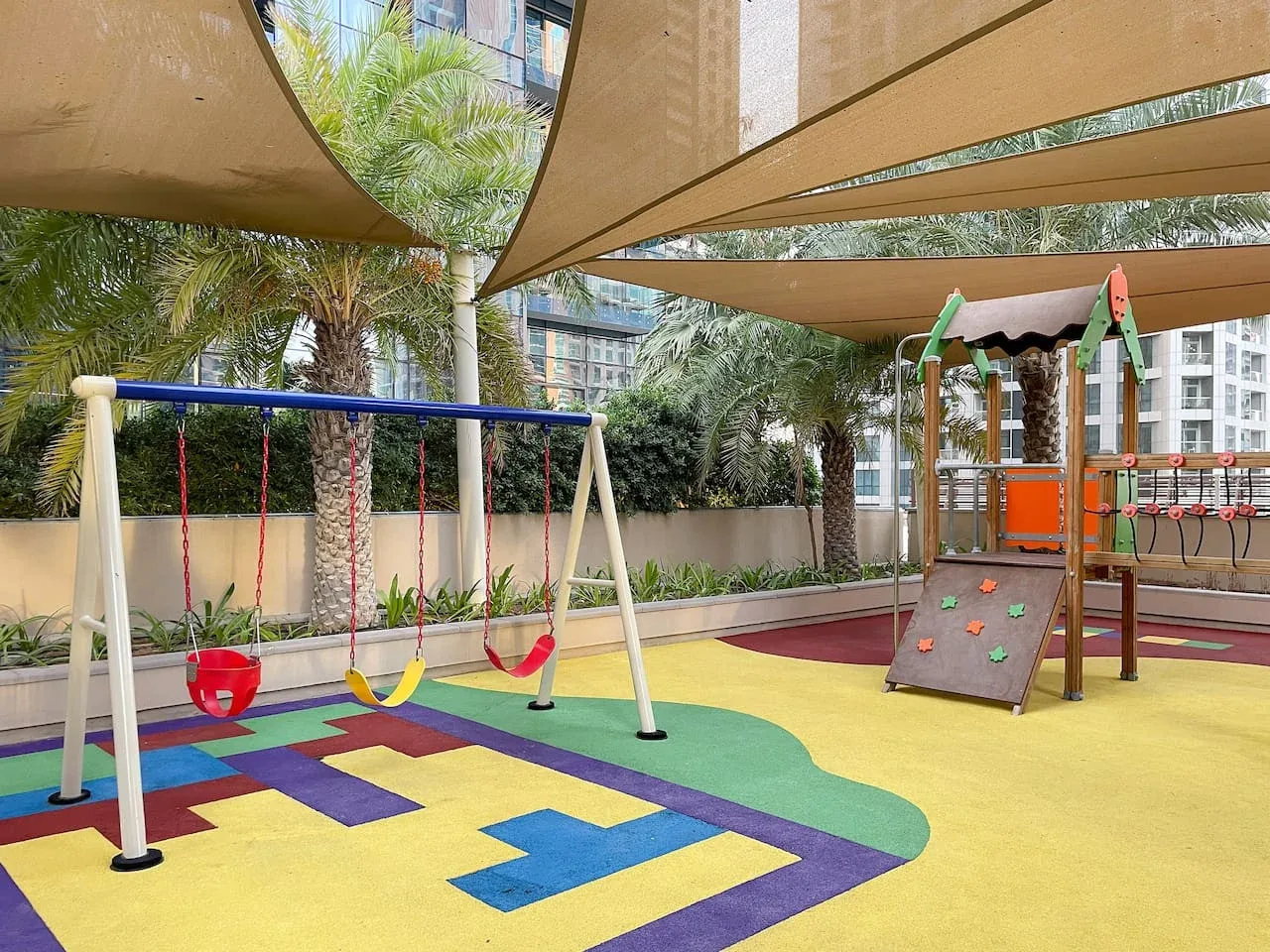 A shaded outdoor playground with swings and a colorful rubber play surface.