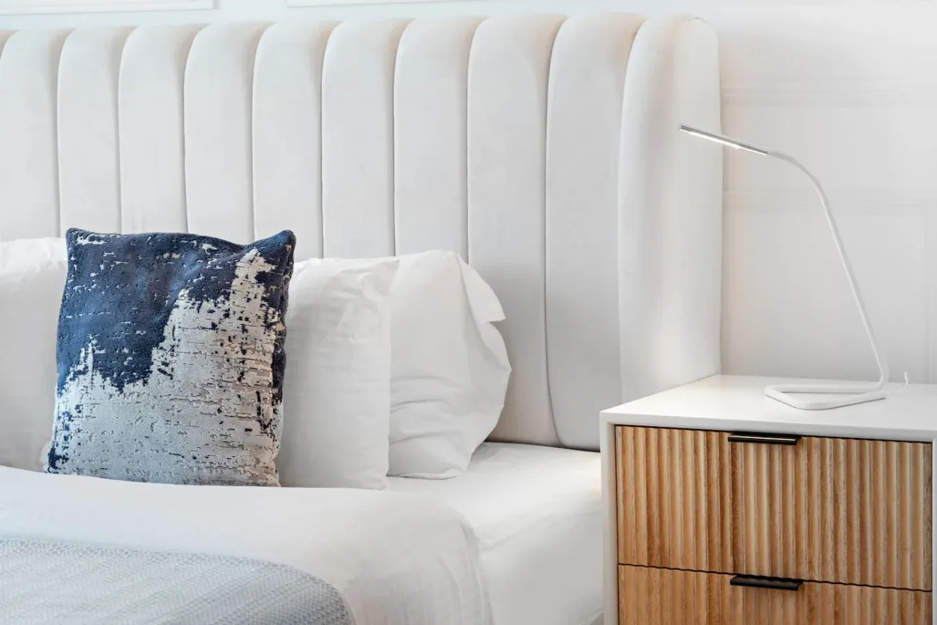 A white bed with a tall upholstered headboard sits beside a wooden nightstand.