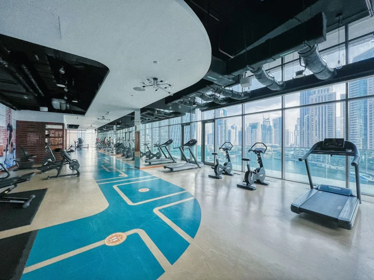 A gym with exercise machines and large windows overlooking city buildings.