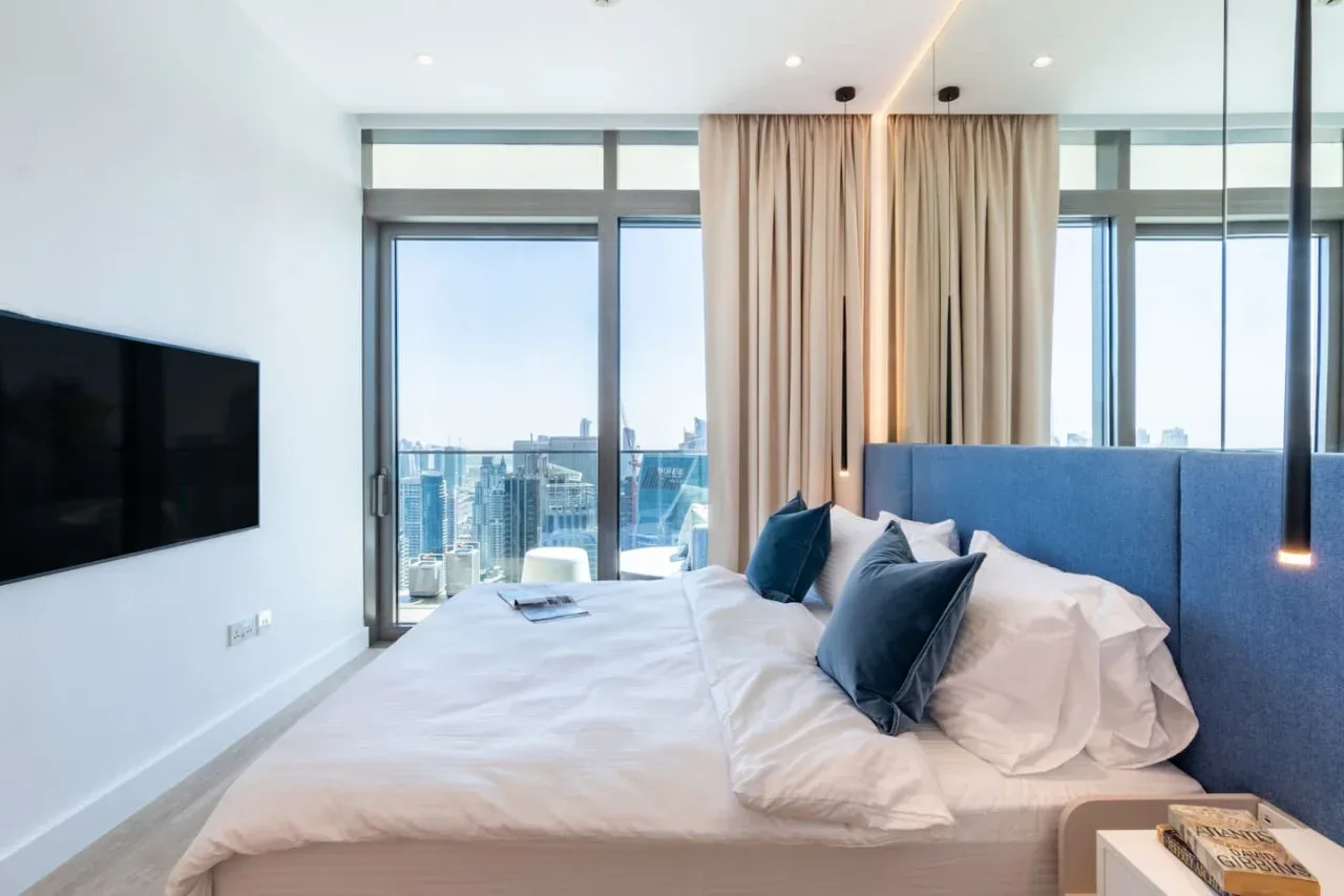 Un dormitorio con cama, cabecero azul y grandes ventanas con vistas a edificios de la ciudad.