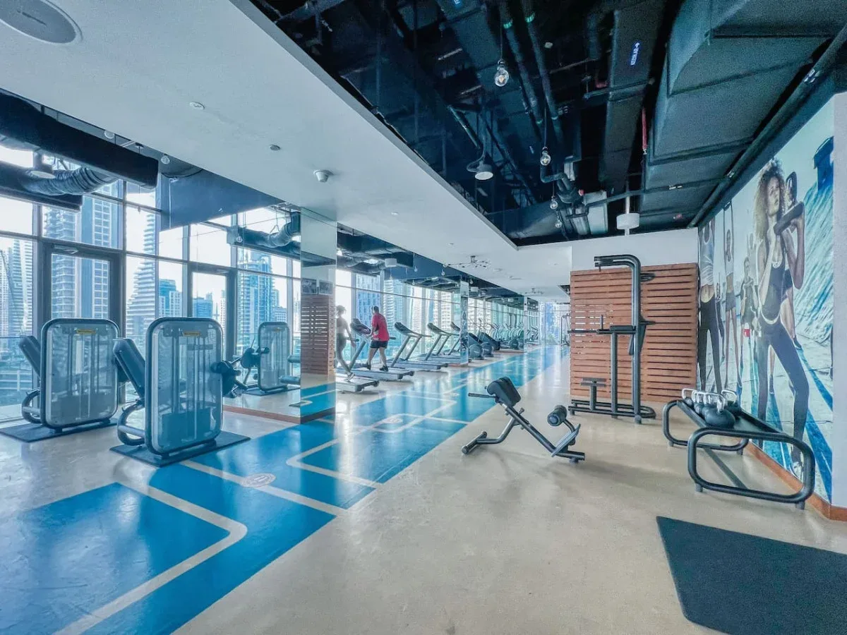 Un gimnasio con máquinas de ejercicio junto a grandes ventanales y un mural colorido.