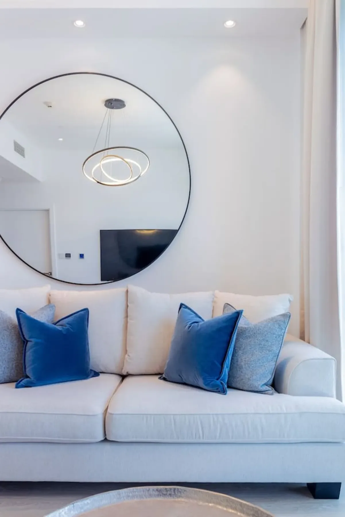 Una sala luminosa con un sofá blanco y cojines azules bajo un gran espejo redondo.