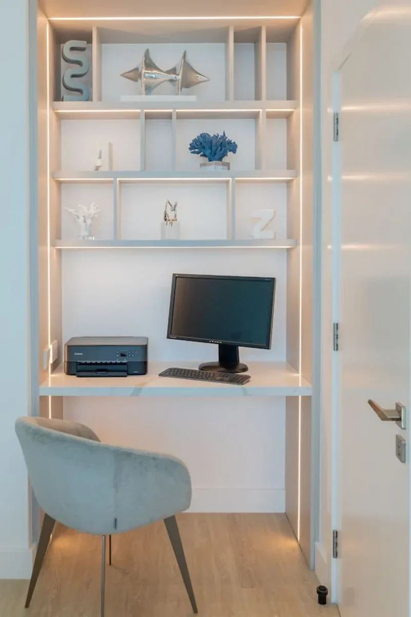 Un rincón de escritorio con computadora, estantes y una silla tapizada azul.
