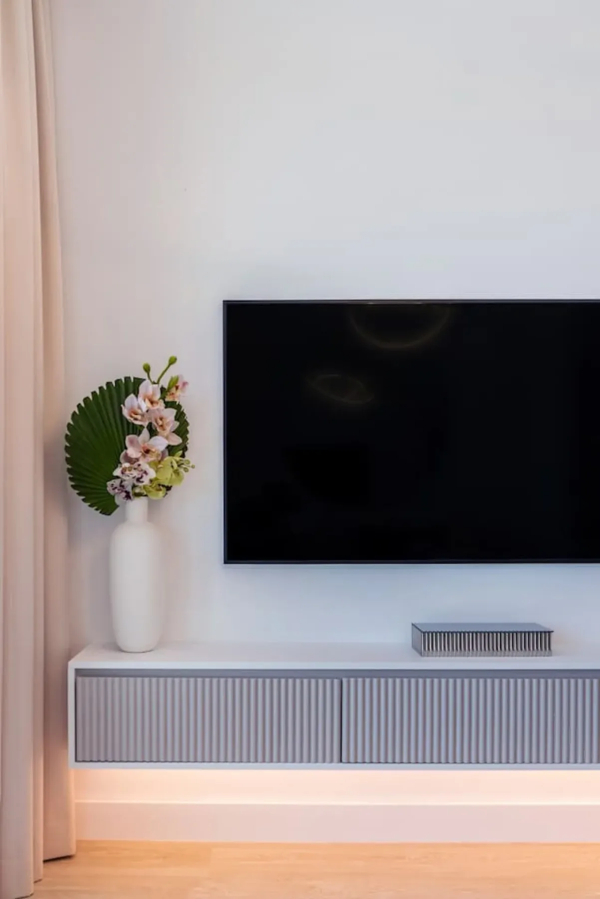 Un televisor cuelga sobre una consola acanalada, con flores al lado.