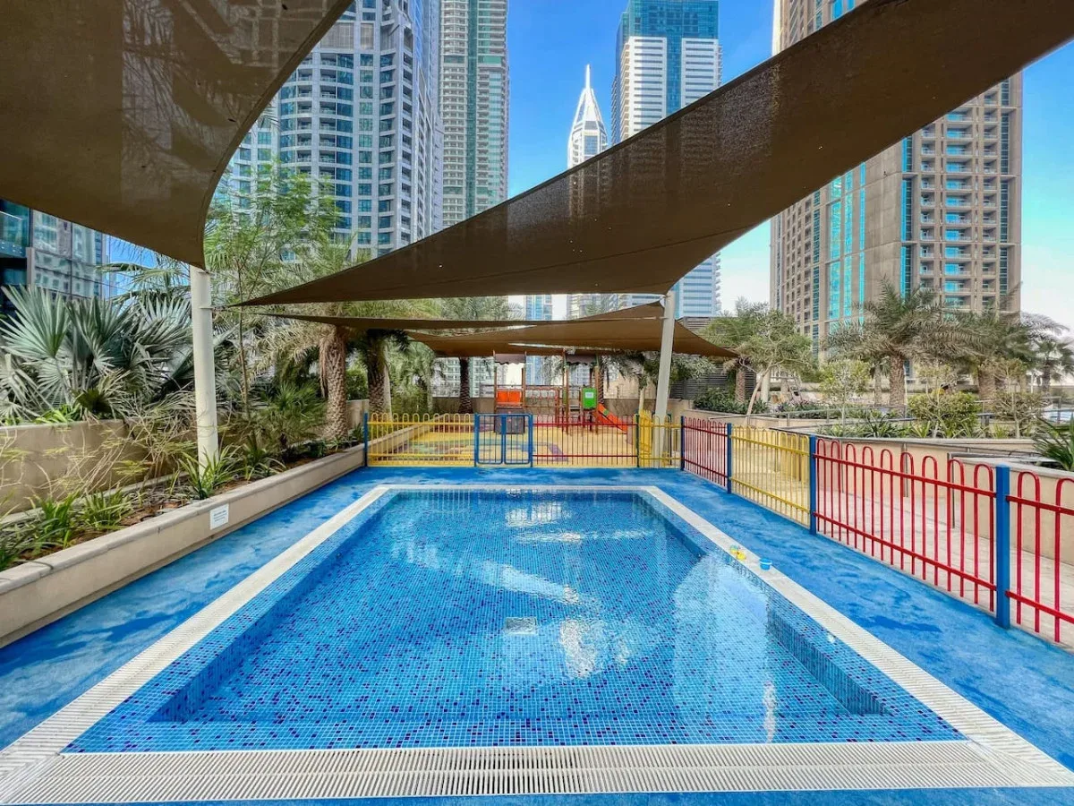 Una piscina azul está bajo toldos de sombra, rodeada de edificios altos.