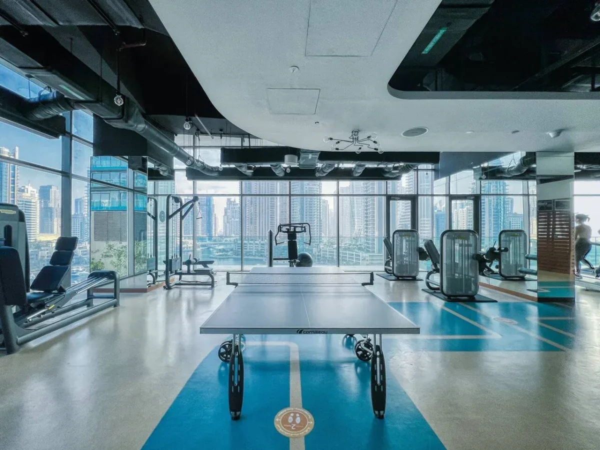 Un gimnasio con equipos de ejercicio y una mesa de tenis de mesa junto a grandes ventanales.