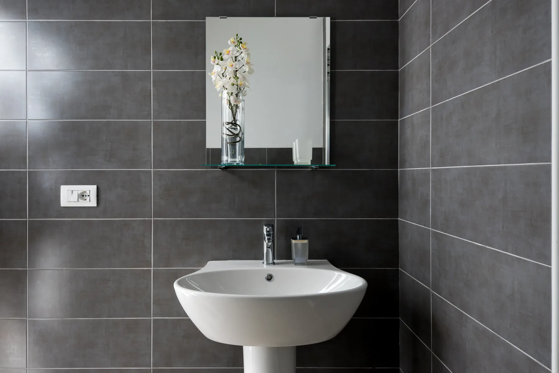 Un lavabo de baño está frente a paredes de azulejos oscuros, con un espejo encima.