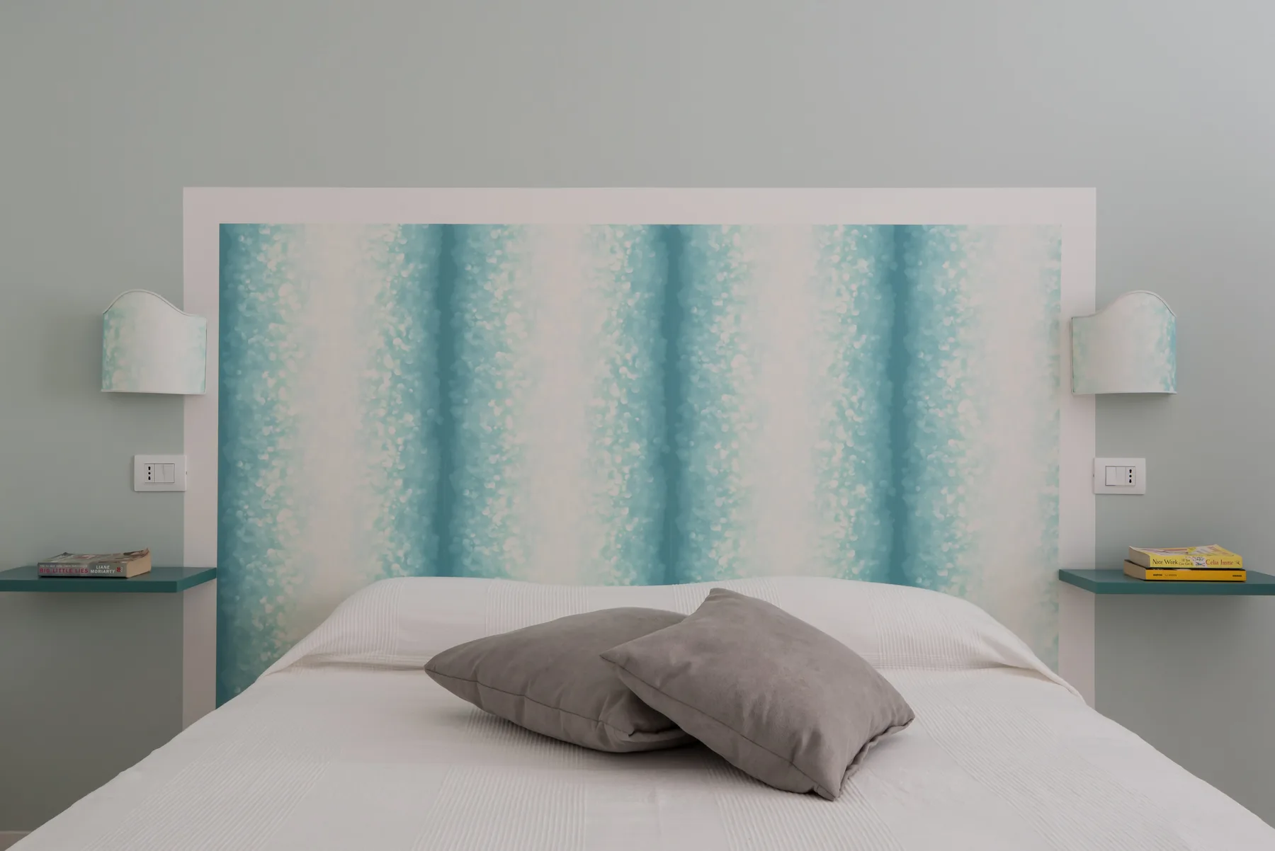 Un dormitorio con una cama y almohadas grises frente a una pared turquesa estampada.