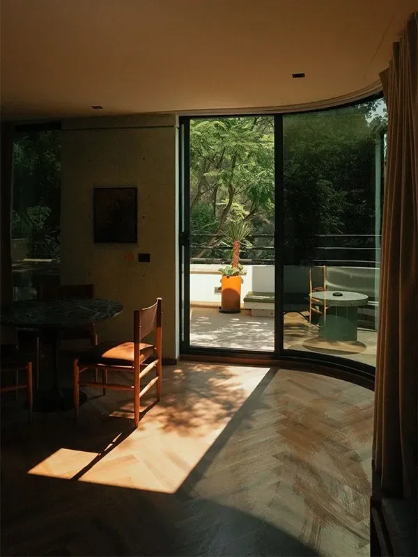 Sunlit interior with wood flooring, a curved glass wall opening toward a balcony with a table and greenery.