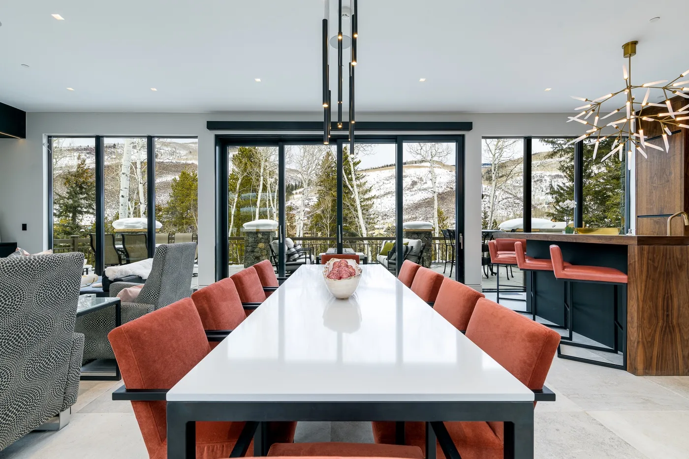 A dining area with a long table and orange seating beside large windows.